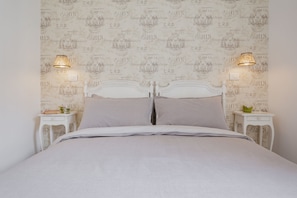 Una cama bien hecha con cabecera blanca, dos mesitas de noche con lámparas y una pared con un motivo repetido que dice 'PARIS' y 'C.R.D'.