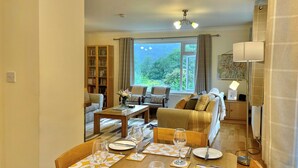 Un coin repas avec une table dressée pour quatre personnes, un canapé, une bibliothèque et une vue sur la verdure à l’extérieur.