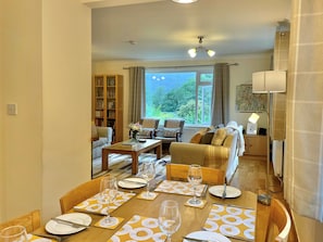 A dining area with a table set for four, a sofa, a bookshelf, and a view of greenery outside.