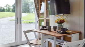 Un coin repas comprenant une table, des chaises, un téléviseur et une vue sur un champ verdoyant, visible à travers une porte vitrée.