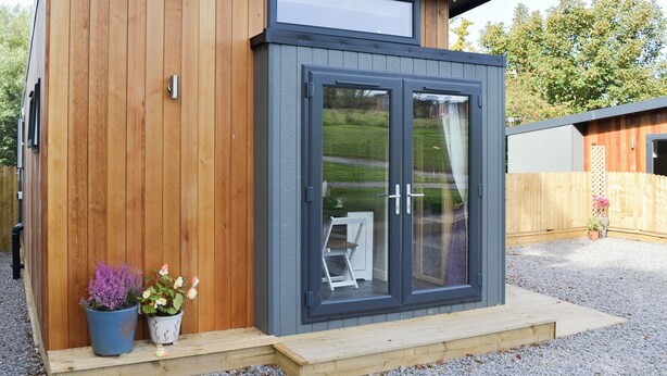 A modern wooden building with a grey door and windows, a small porch, and potted plants. - 0 bedroom accommodation in Brocklebank, near Caldbeck and Uldale (Brocklebank, near Caldbeck and Uldale)