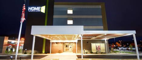 L’entrée d’un hôtel, avec un passage couvert, un drapeau américain et une enseigne indiquant « HOME SUITES BY MILTON ».