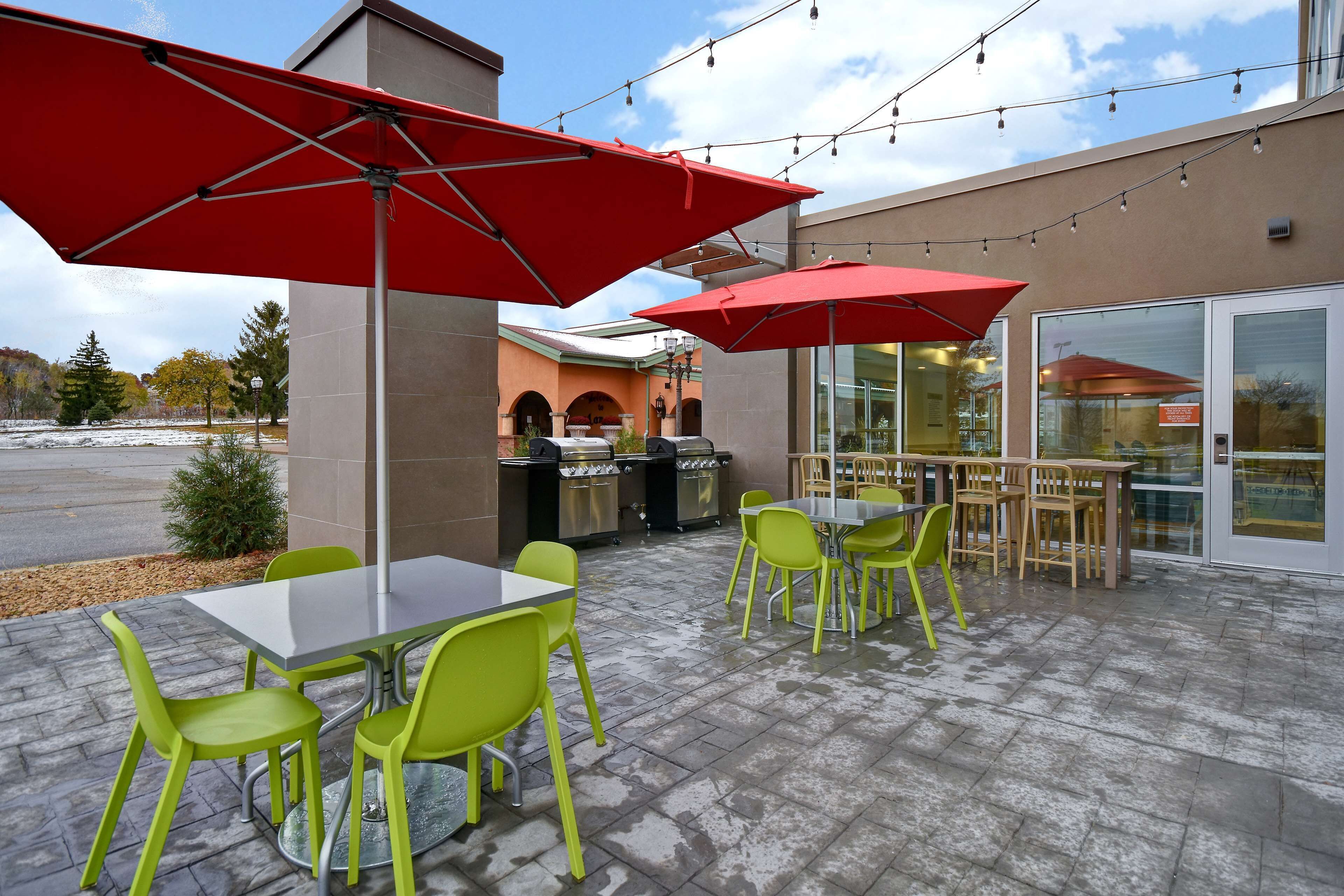 An outdoor patio area with tables and chairs, red umbrellas, and a building with glass doors in the background.