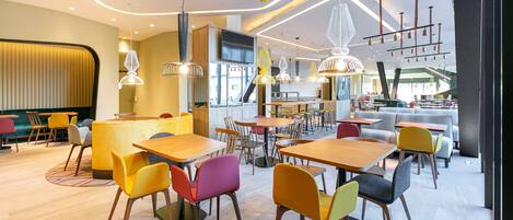 A modern restaurant with wooden tables and colorful chairs, a bar area, and a large window overlooking the city.