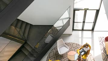 Un interior moderno con una escalera, un área de descanso con mesas redondas y una barandilla de vidrio.