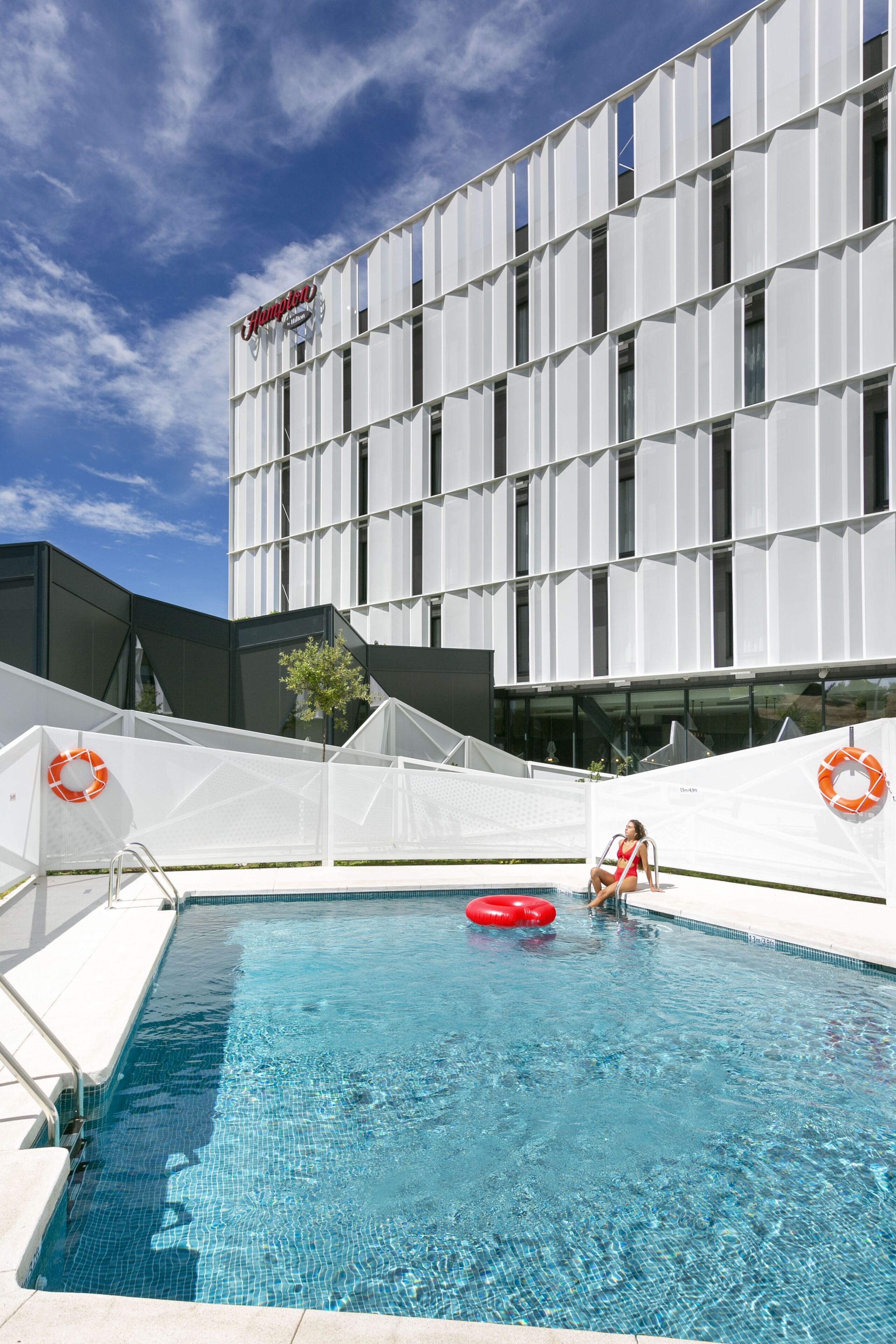 Una piscina con una persona en traje de baño rojo, un flotador salvavidas y un edificio con un letrero al fondo.