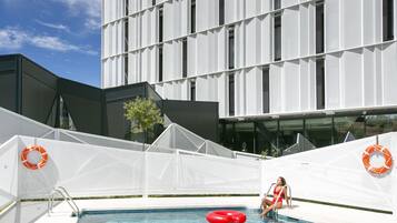 Una piscina con una persona en traje de baño rojo, un flotador salvavidas y un edificio con un letrero al fondo.