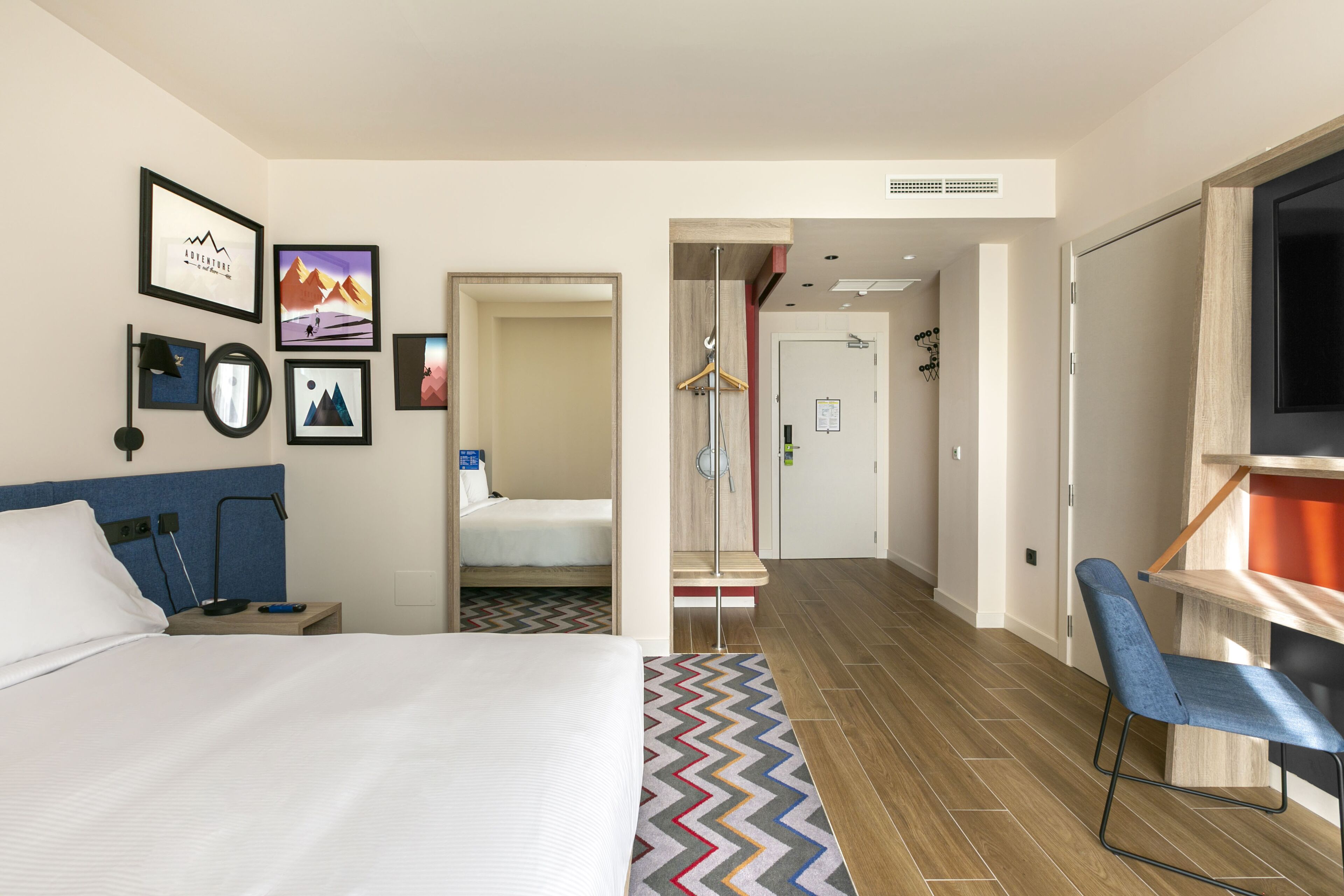 A hotel room with a bed, a desk, a chair, a TV, and a bathroom visible through an open door.