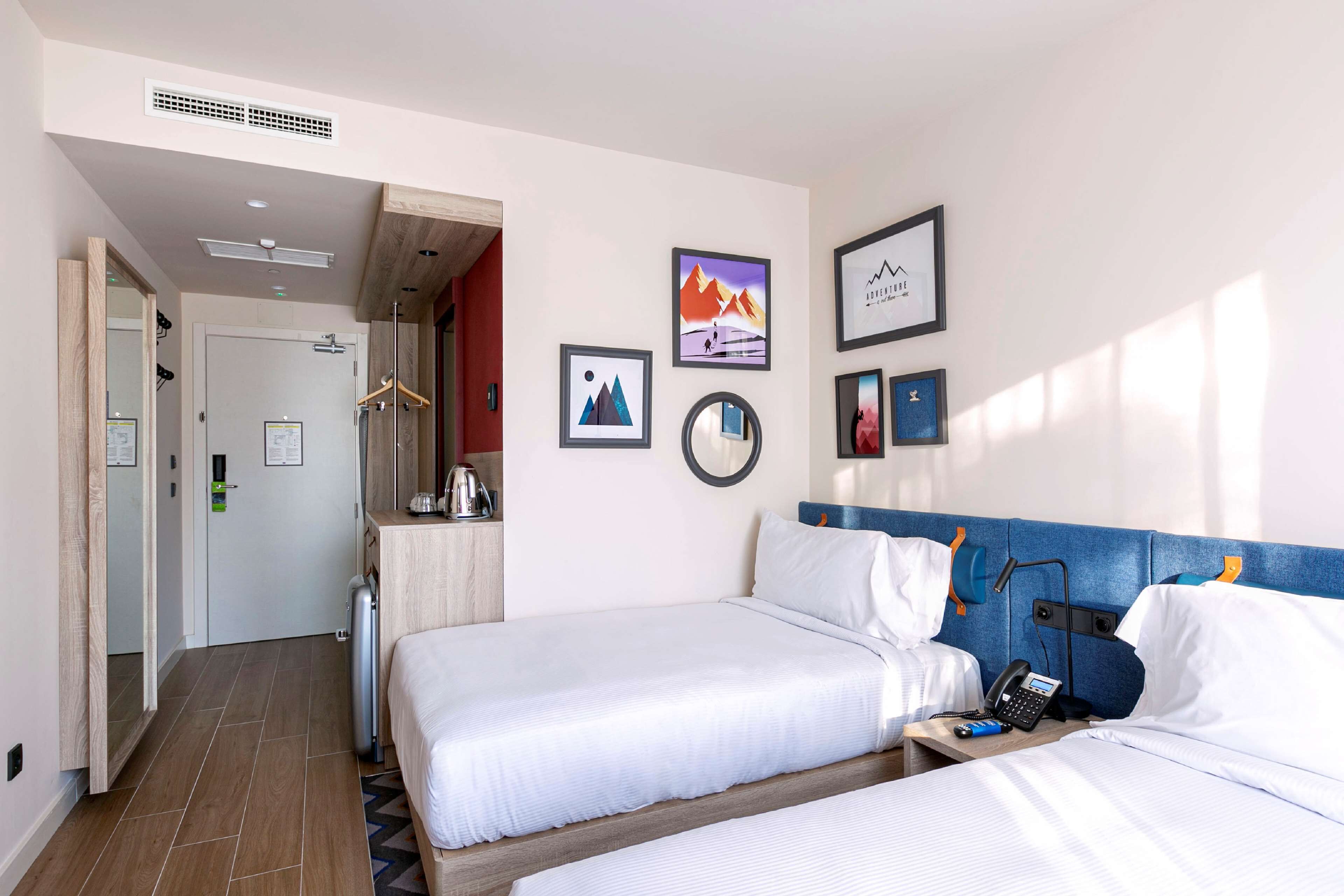 A hotel room with two beds, a desk with a phone, and a mirror. There are framed pictures on the wall and a door leading to another room.