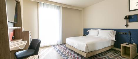Una habitación de hotel con cama, escritorio, silla y un ventanal con cortinas.