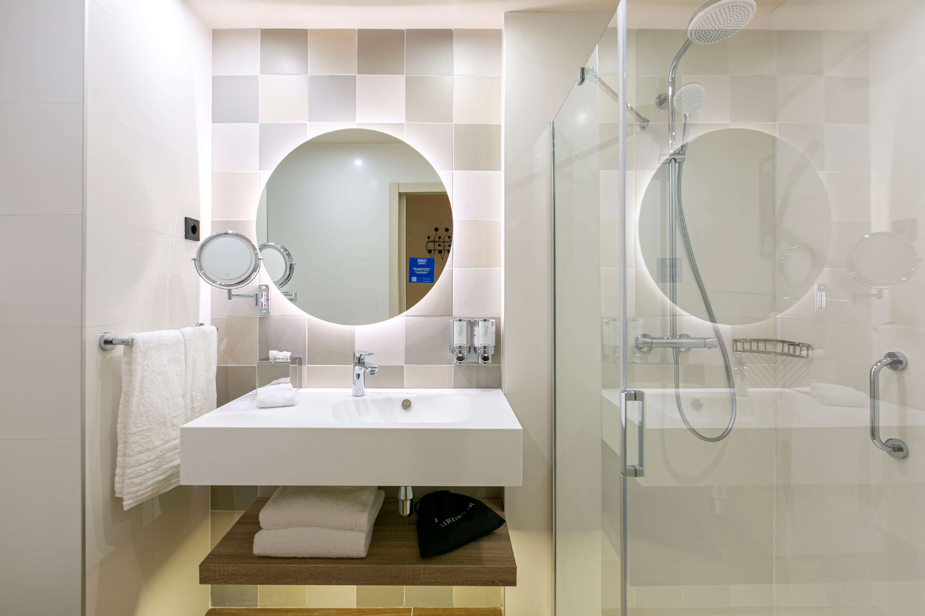 Une salle de bain moderne avec un lavabo blanc, un miroir rond et une douche à parois de verre.