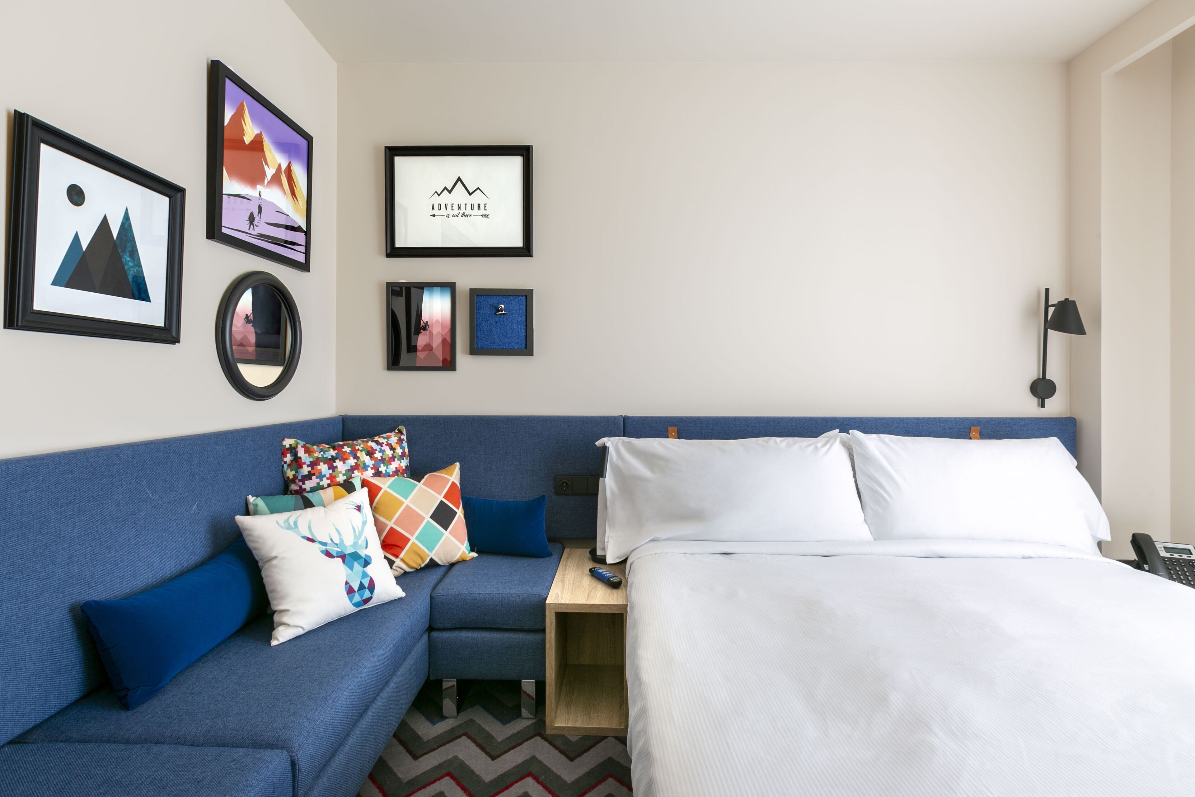 Quarto de hotel com sofá azul, cama com roupa de cama branca e uma pequena mesa de cabeceira de madeira. Há quadros nas paredes e uma luminária na mesinha de apoio.