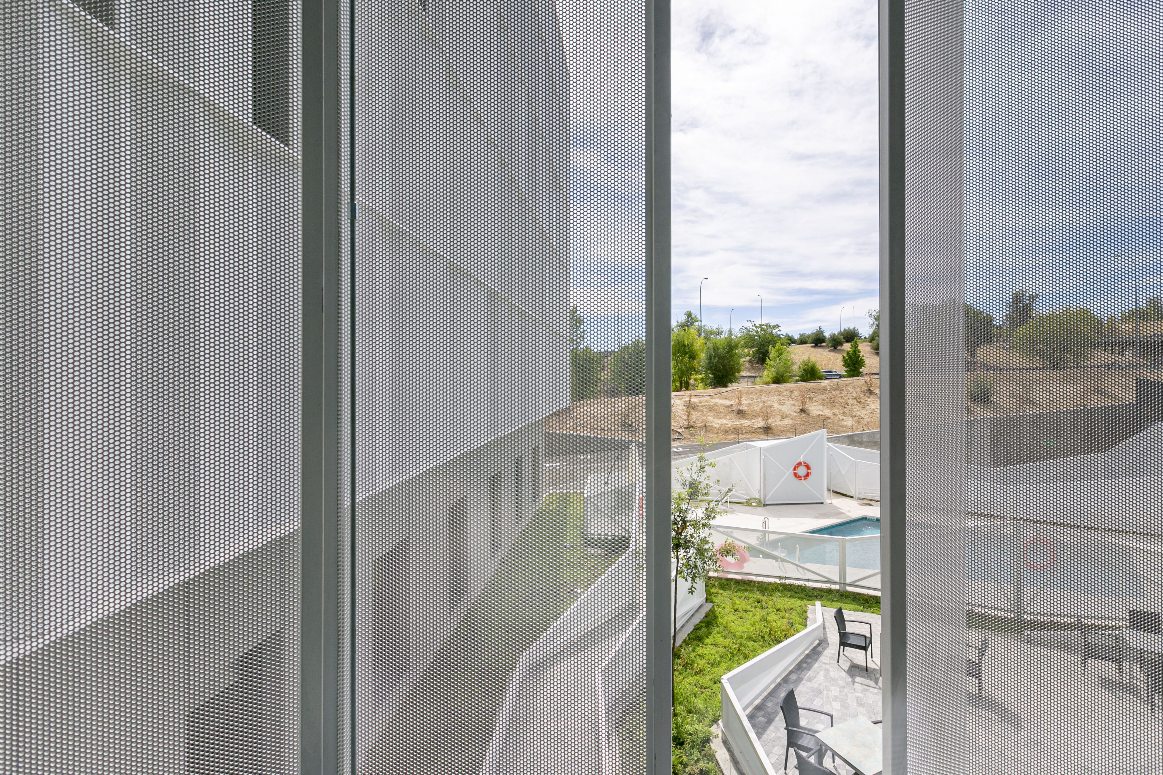 A view through a window with a mesh screen, revealing a pool area with outdoor seating and a landscaped garden.