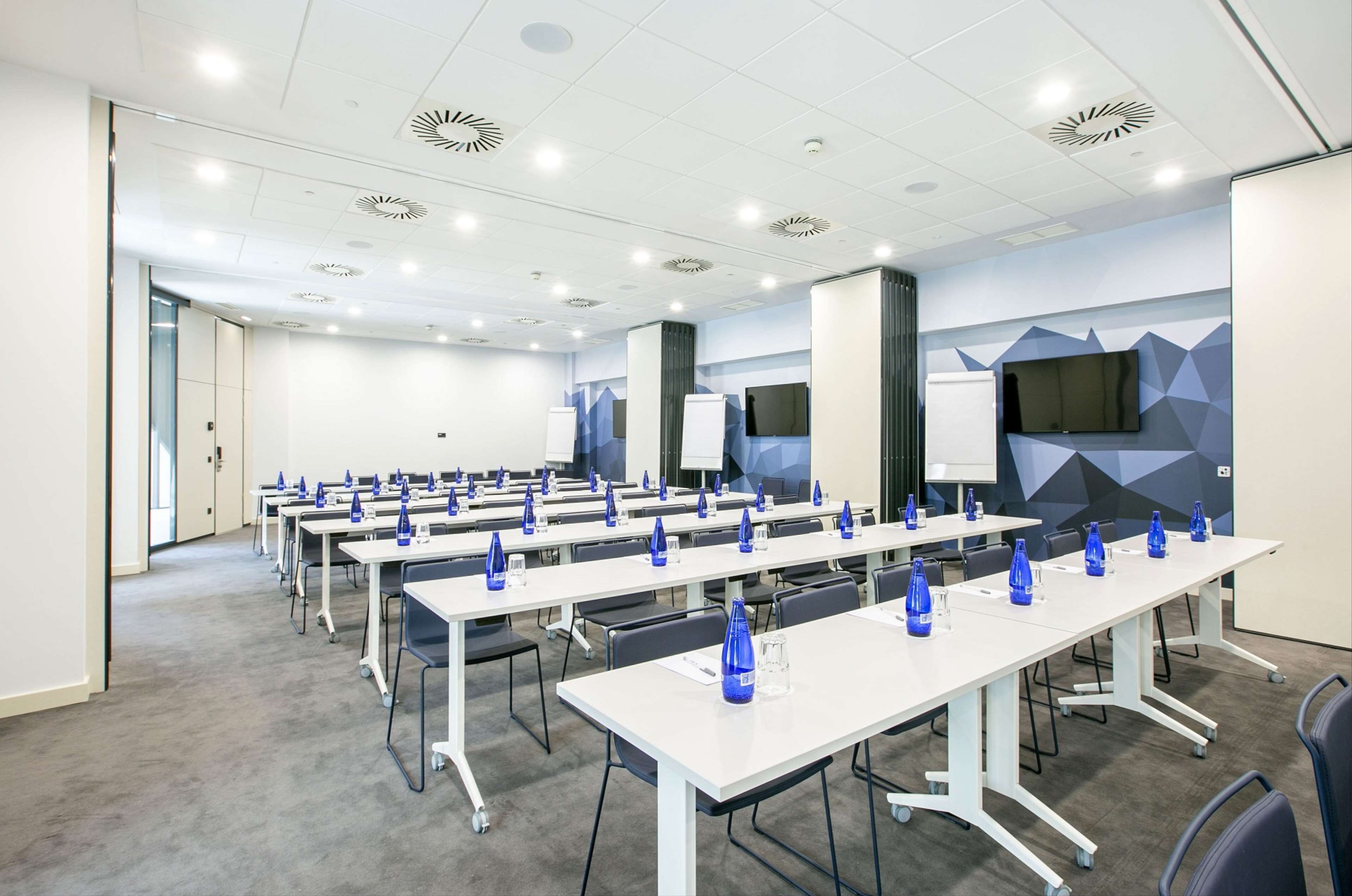 Une salle de conférence avec plusieurs tables blanches, des bouteilles d’eau bleues et un grand écran mural.