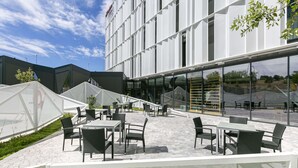 An outdoor seating area with tables and chairs, a modern building with large windows, and a clear sky. - Hampton by Hilton Alcobendas Madrid (Alcobendas)