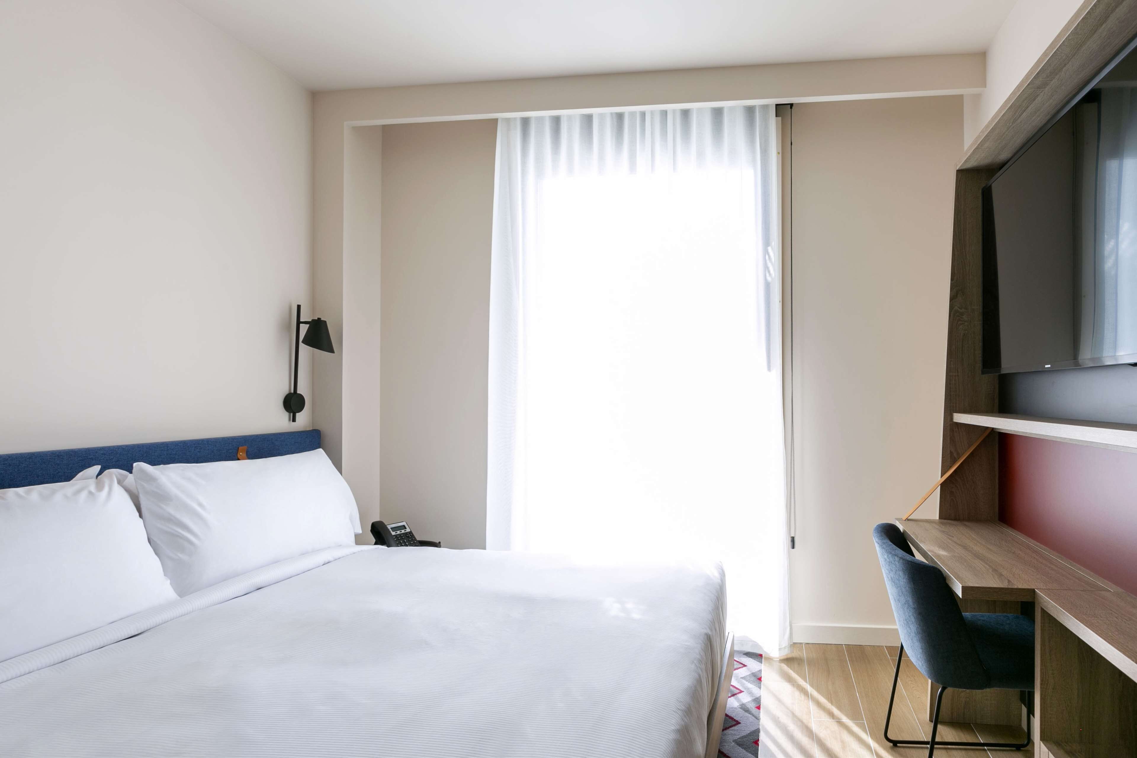 A hotel room with a bed, a desk, a chair, a TV, and a window with curtains.