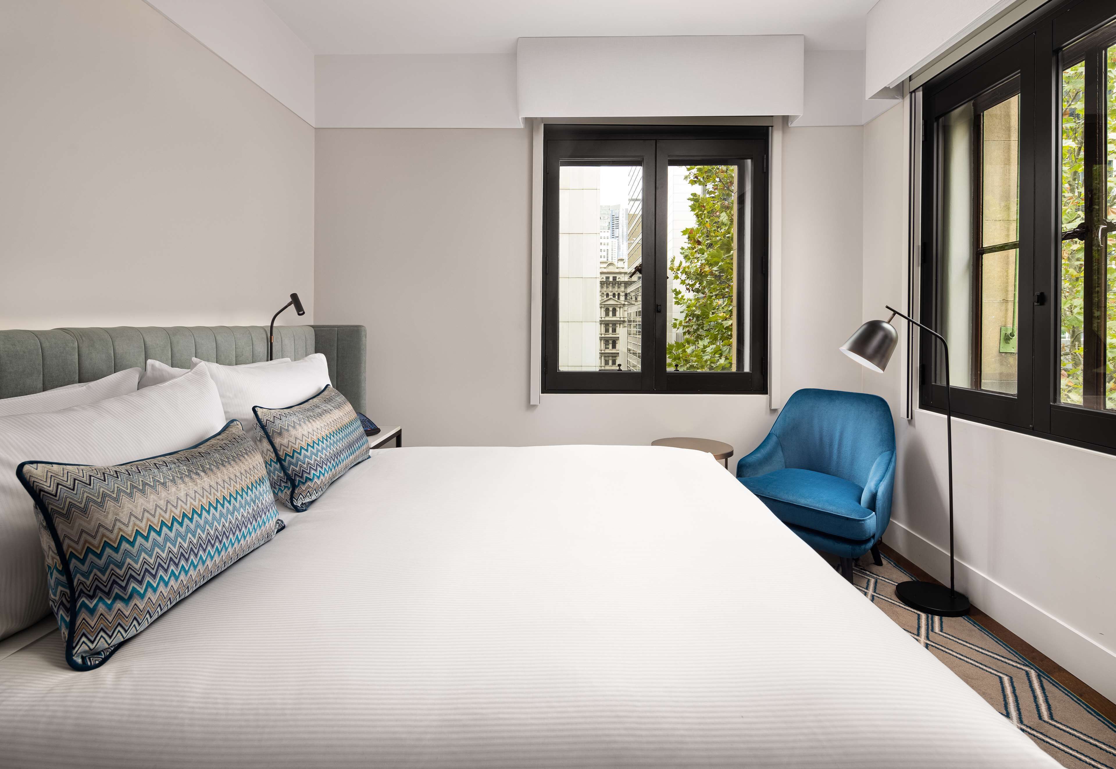 A modern hotel room with a large bed, a blue armchair, and a window with a city view.