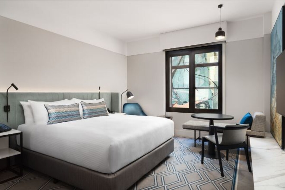 A modern hotel room with a large bed, a small table, a chair, and a window with a view.