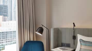 A hotel room with a large bed, a blue armchair, a bedside table with a lamp, and a view of the city through the window.