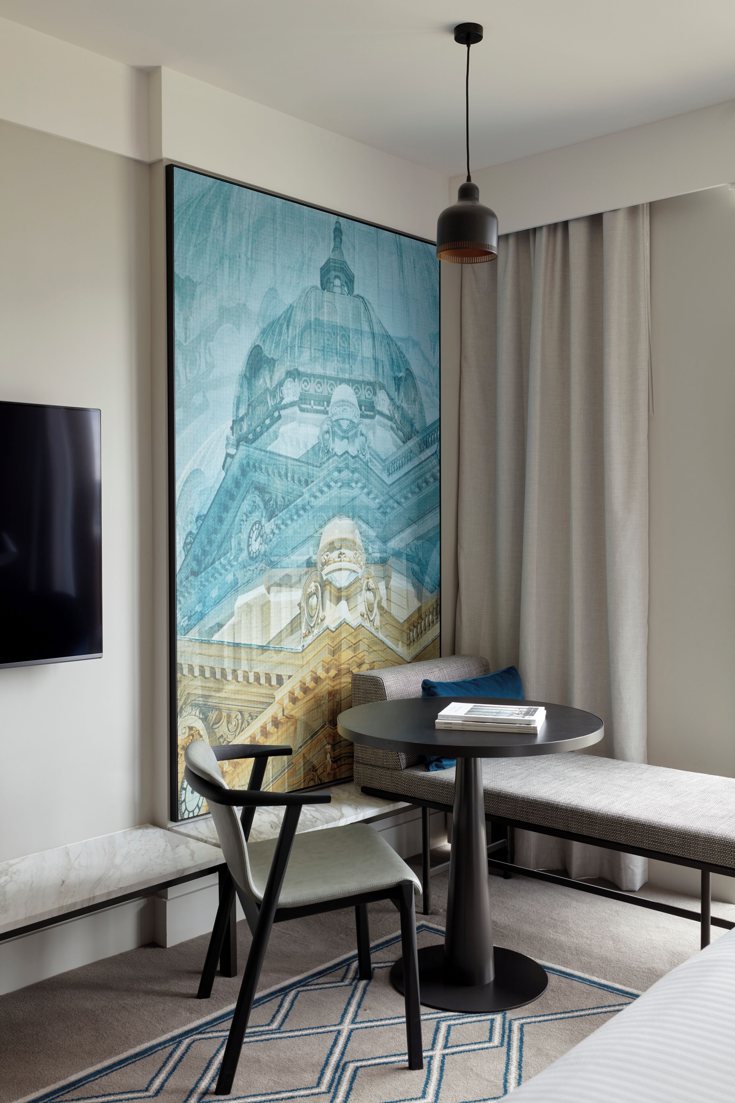 Une chambre d’hôtel moderne avec une grande fresque murale, une table ronde avec une chaise, un téléviseur fixé au mur et un luminaire suspendu.