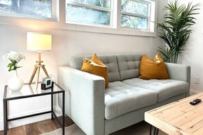 Ein Wohnzimmer mit grauem Sofa, zwei gelben Kissen, einem kleinen Beistelltisch mit Lampe, einer Vase mit Blumen und einer großen Pflanze.