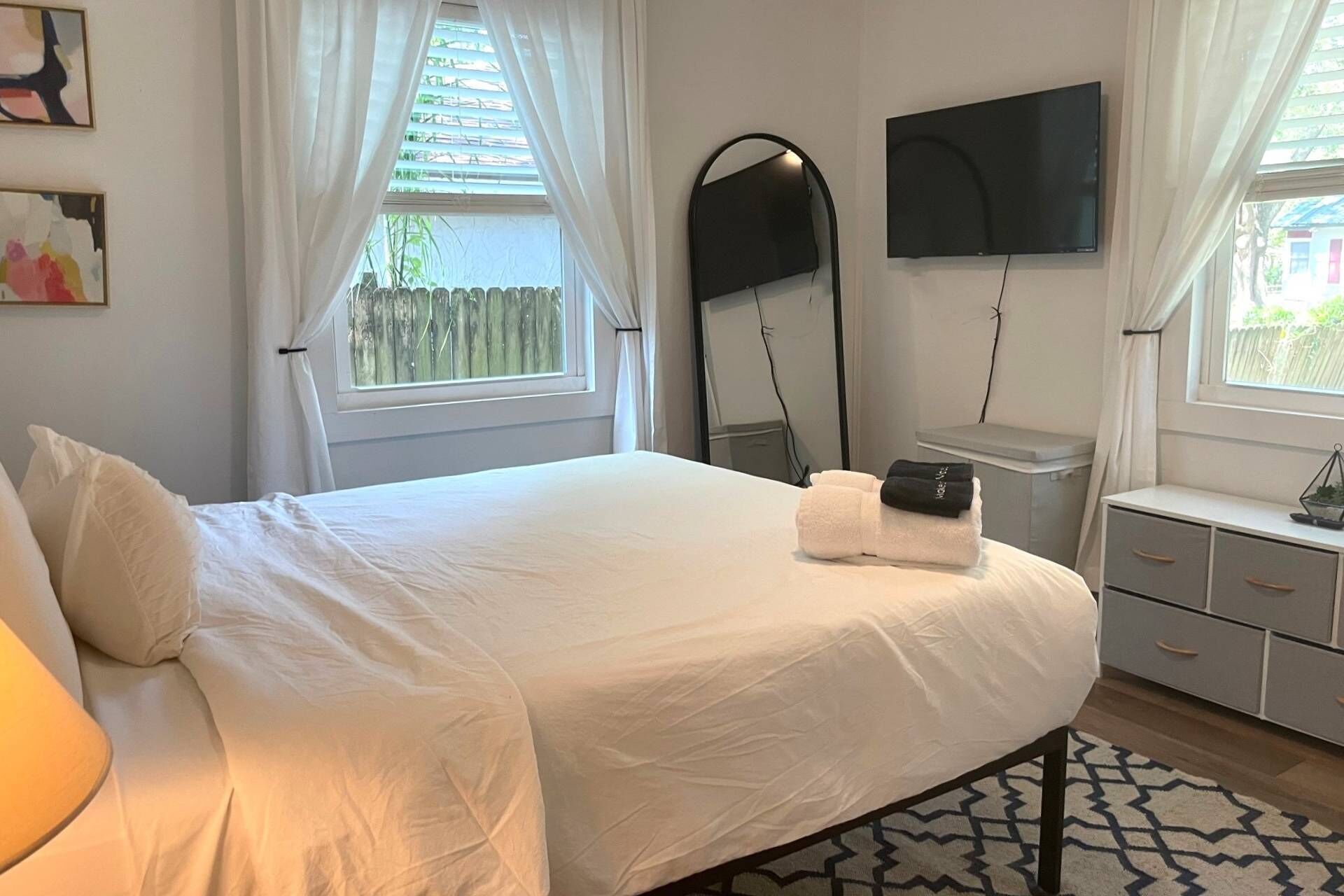 Een net opgemaakt bed met witte lakens, een nachtkastje met een lamp, een flatscreen televisie aan de muur en een raam met witte gordijnen.