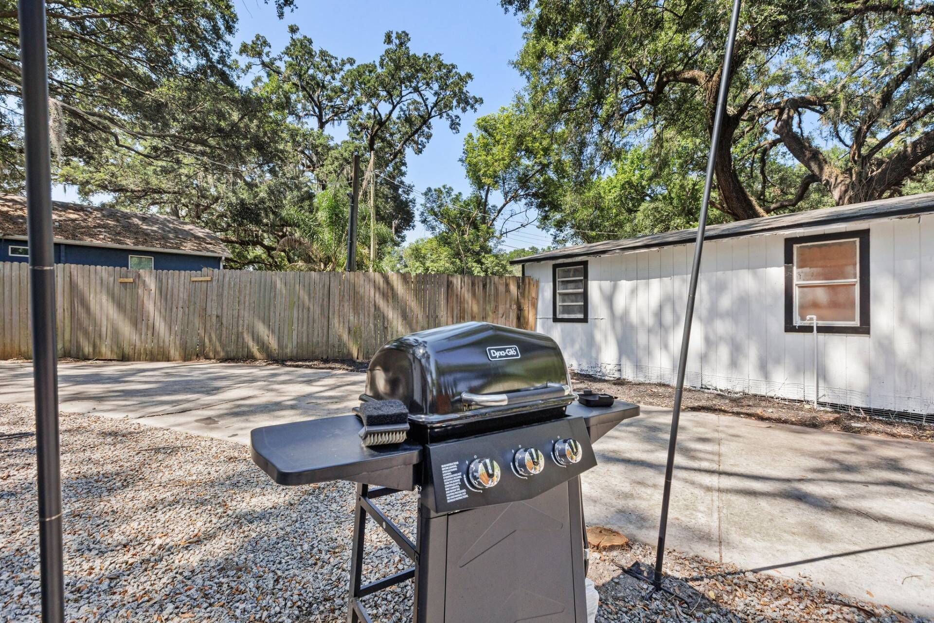 A portable grill with a digital display and control knobs, positioned on a gravel surface in a backyard with a wooden fence and a small shed in the background.