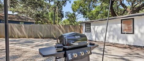 A portable grill with a digital display and control knobs, positioned on a gravel surface in a backyard with a wooden fence and a small shed in the background.