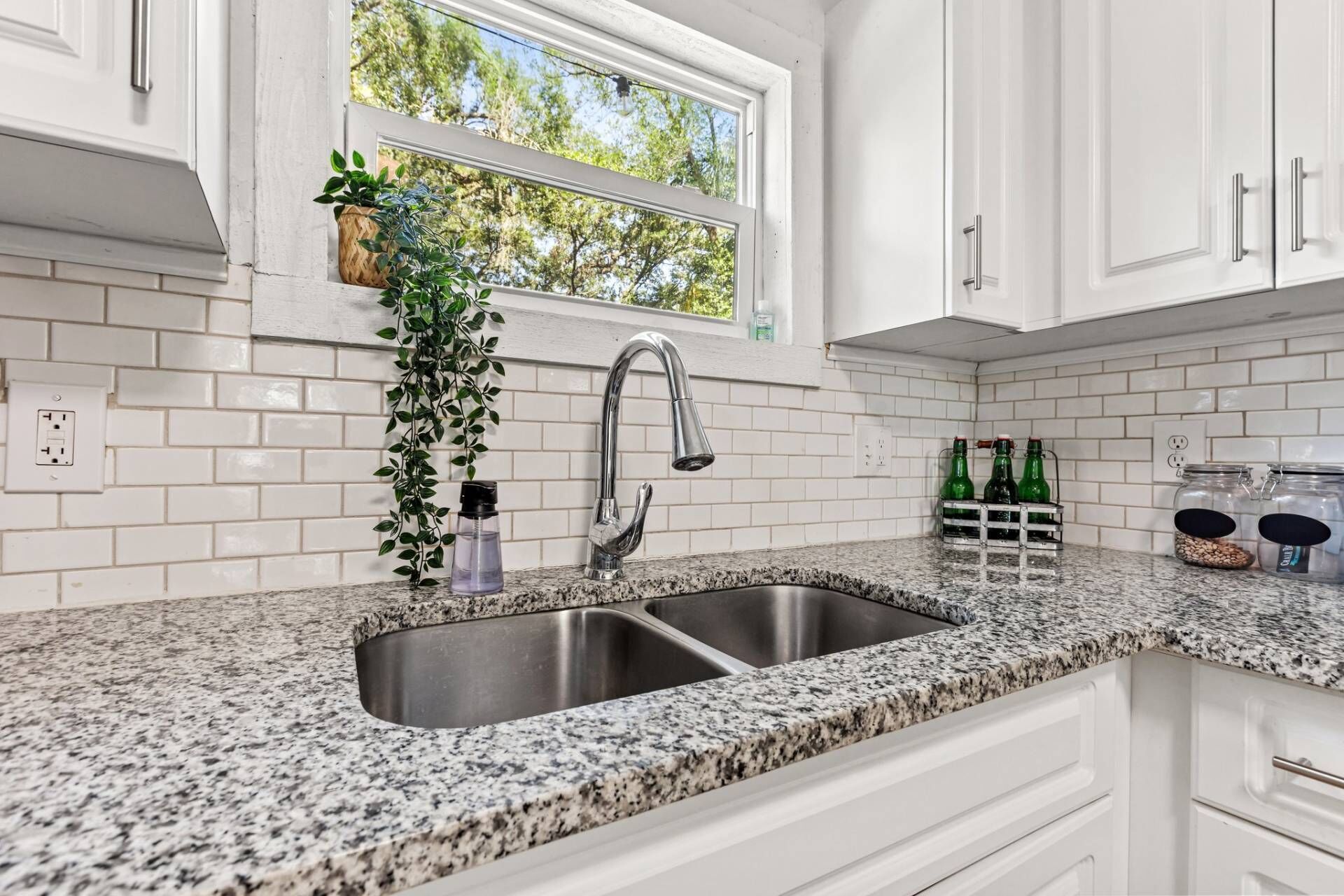 A kitchen with white cabinets, a granite countertop, a stainless steel sink, a window with a view of trees, and a plant hanging above the sink.
