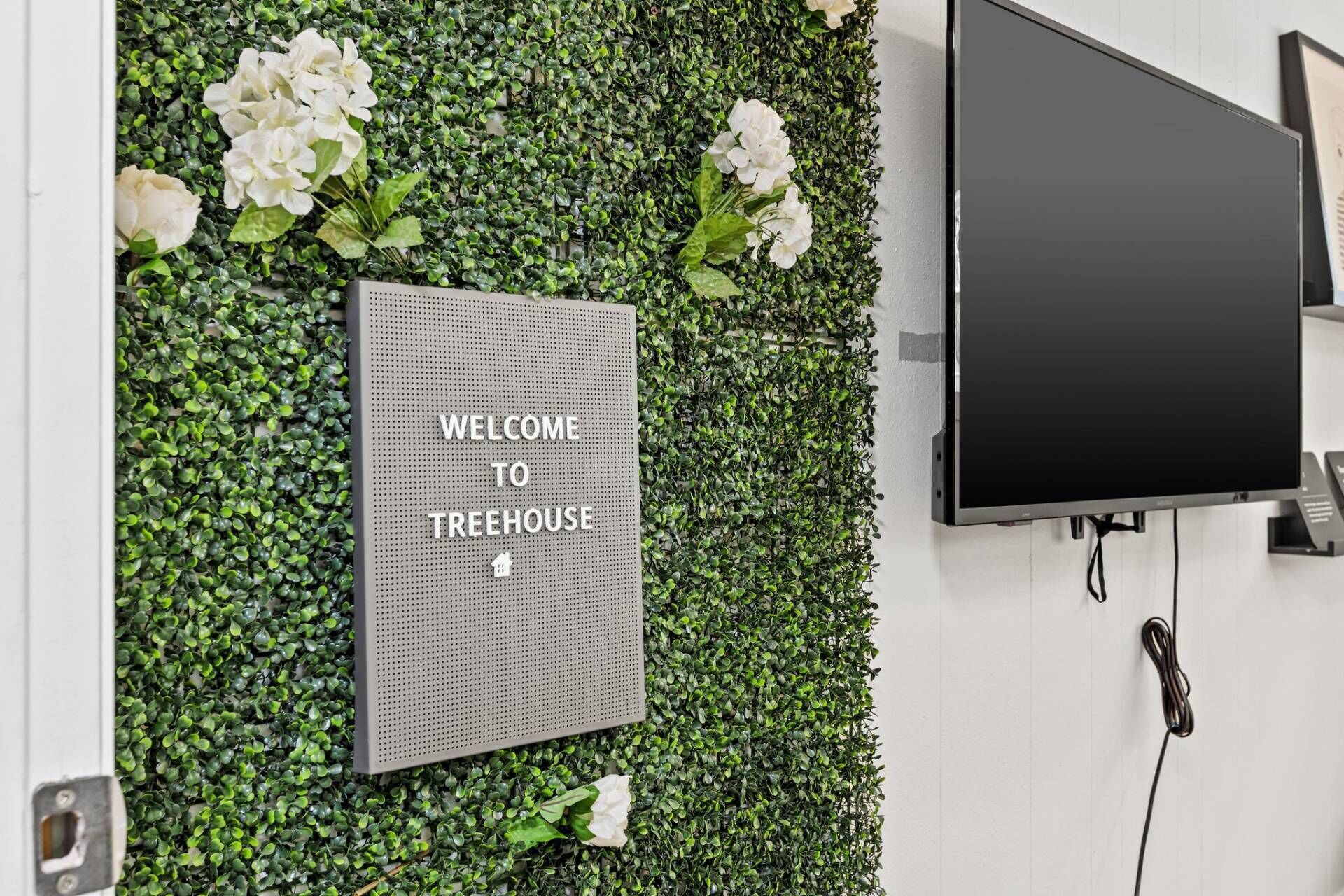 A welcome sign on a wall with a green foliage backdrop and a flat-screen TV mounted on the wall.