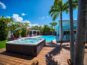 A wooden deck with a hot tub, a swimming pool, and a covered patio area with chairs and umbrellas.