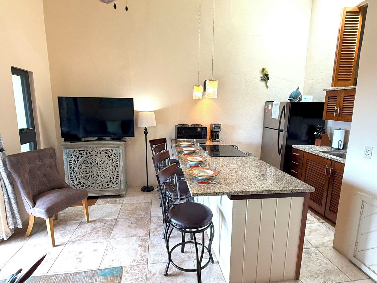 A modern kitchen with a central island, bar stools, a flat-screen TV, and a dining area with chairs.