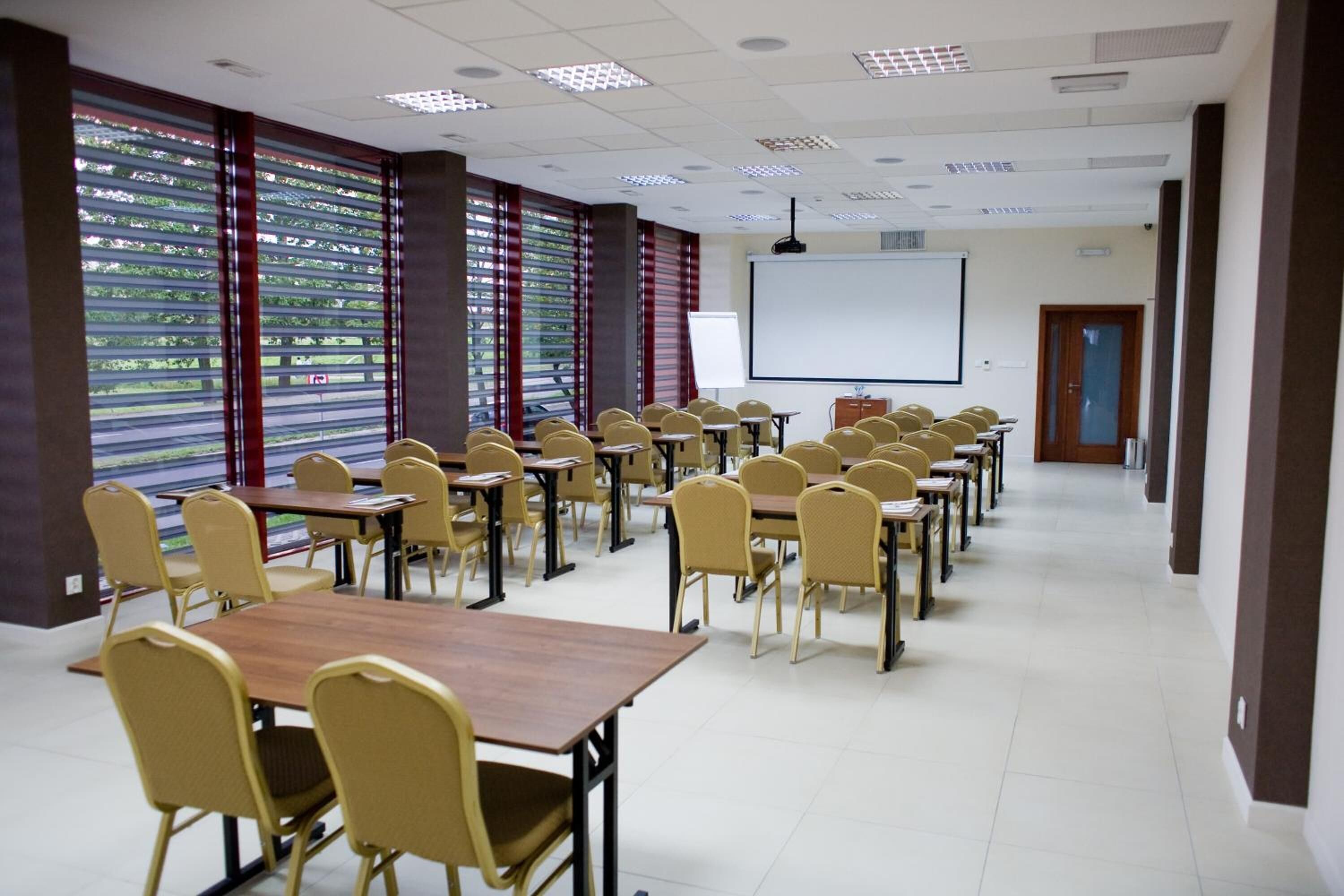Un salón de conferencias con filas de mesas y sillas, una pantalla de proyector y un ventanal con persianas.