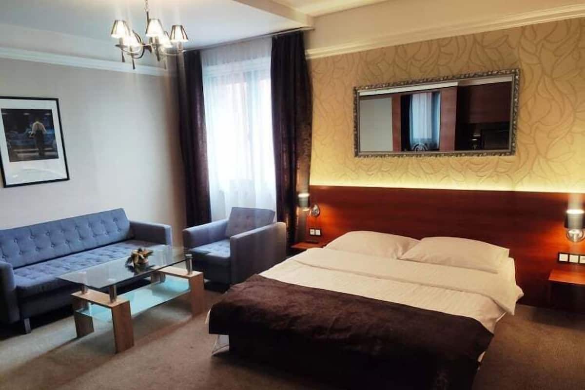 A hotel room with a bed, a sofa, a chair, a coffee table, a chandelier, a large mirror, and a framed picture on the wall.
