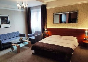A hotel room with a bed, a sofa, a chair, a coffee table, a chandelier, a large mirror, and a framed picture on the wall. - Hotel & Spa Kameleon (Zory)