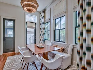 A modern living room with a wooden dining table, white chairs, a sofa, and large windows with curtains.
