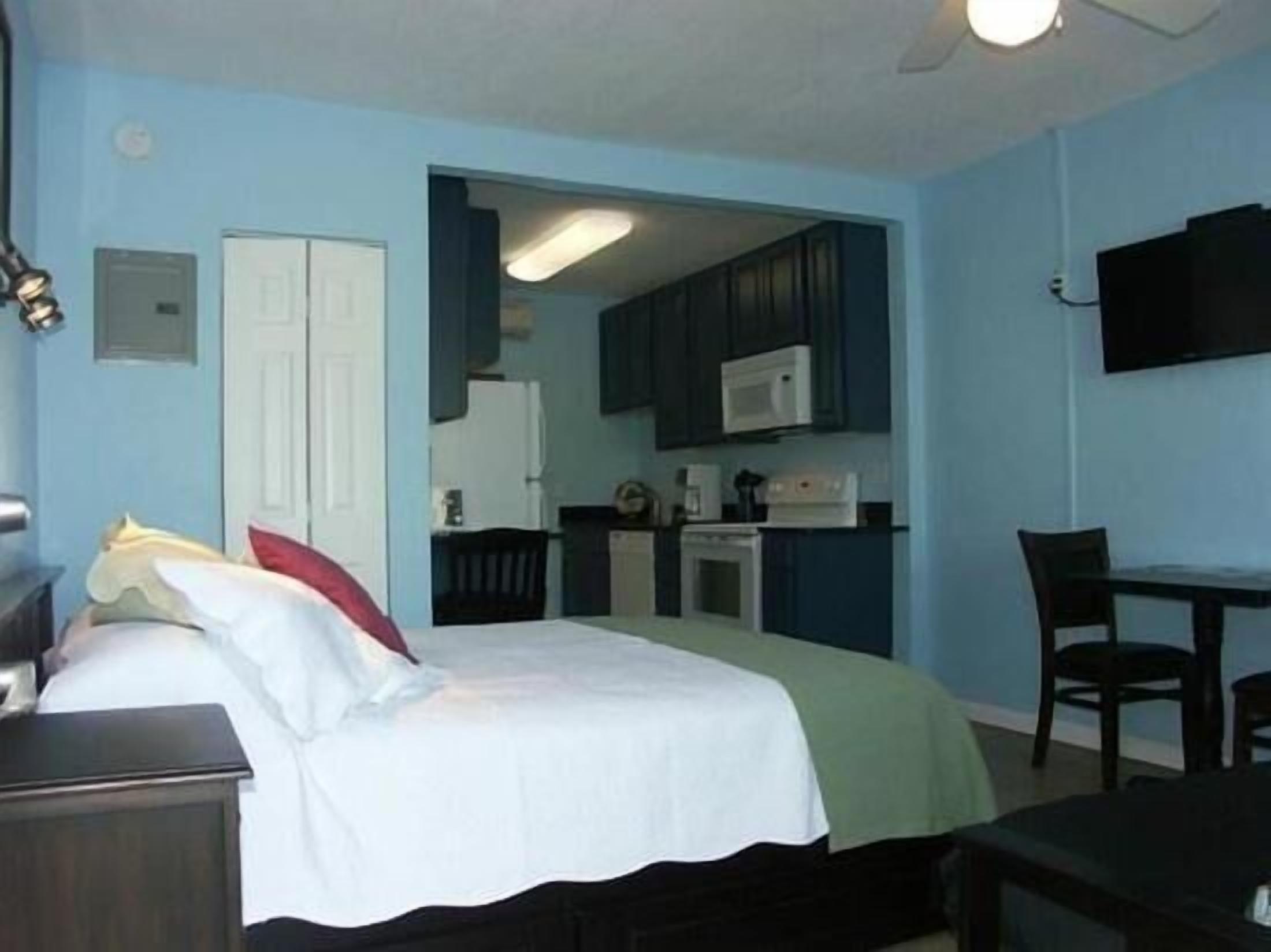 A compact hotel room with a bed, a small desk, a chair, a kitchenette, and a TV.