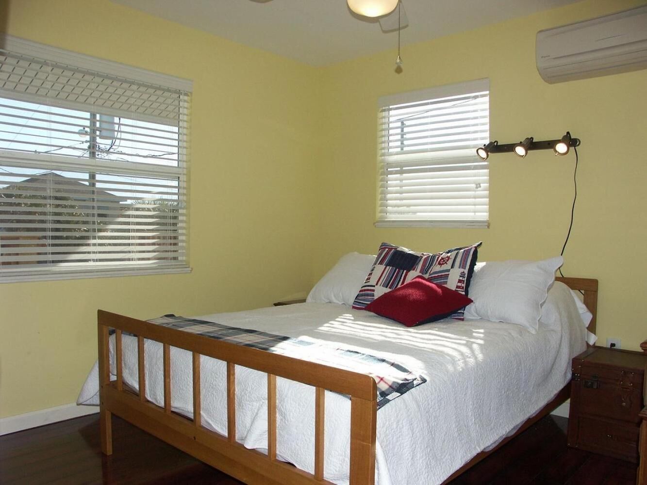 Un dormitorio con una cama de madera, ropa de cama blanca y una almohada roja. Hay dos ventanas con persianas, un ventilador de techo y un aire acondicionado.
