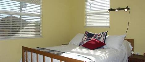 Un dormitorio con una cama de madera, ropa de cama blanca y una almohada roja. Hay dos ventanas con persianas, un ventilador de techo y un aire acondicionado.