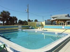 A rectangular swimming pool with a blue and white tiled edge, surrounded by a yellow and blue fence. There are lounge chairs and a small table near the pool. - Flagler Beach Motel and Vacation Rentals (Flagler Beach)