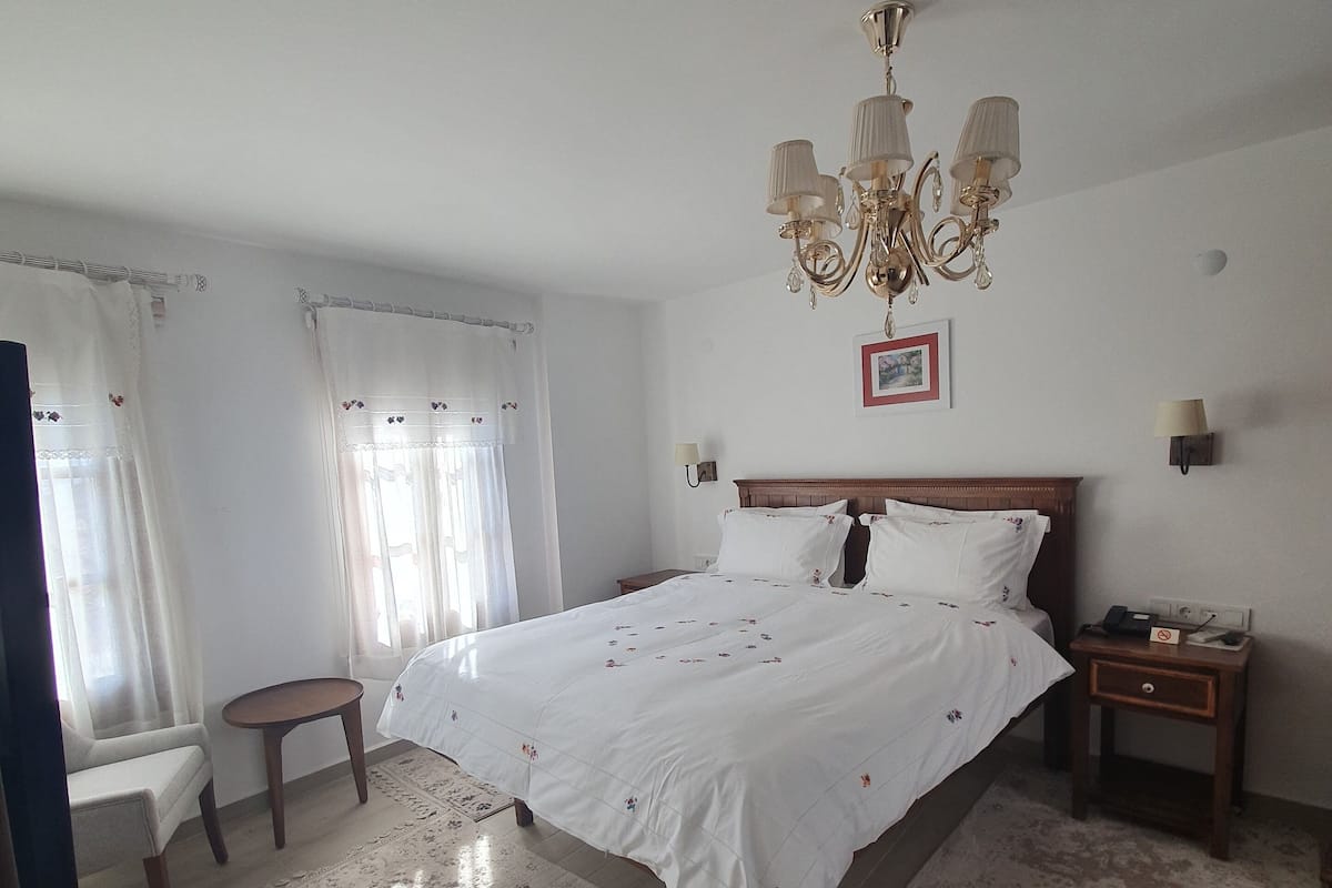 Ein Schlafzimmer mit einem großen Bett, einer Kandelaber, einem Nachttisch, einem kleinen Tisch und einem Fenster mit Vorhängen.