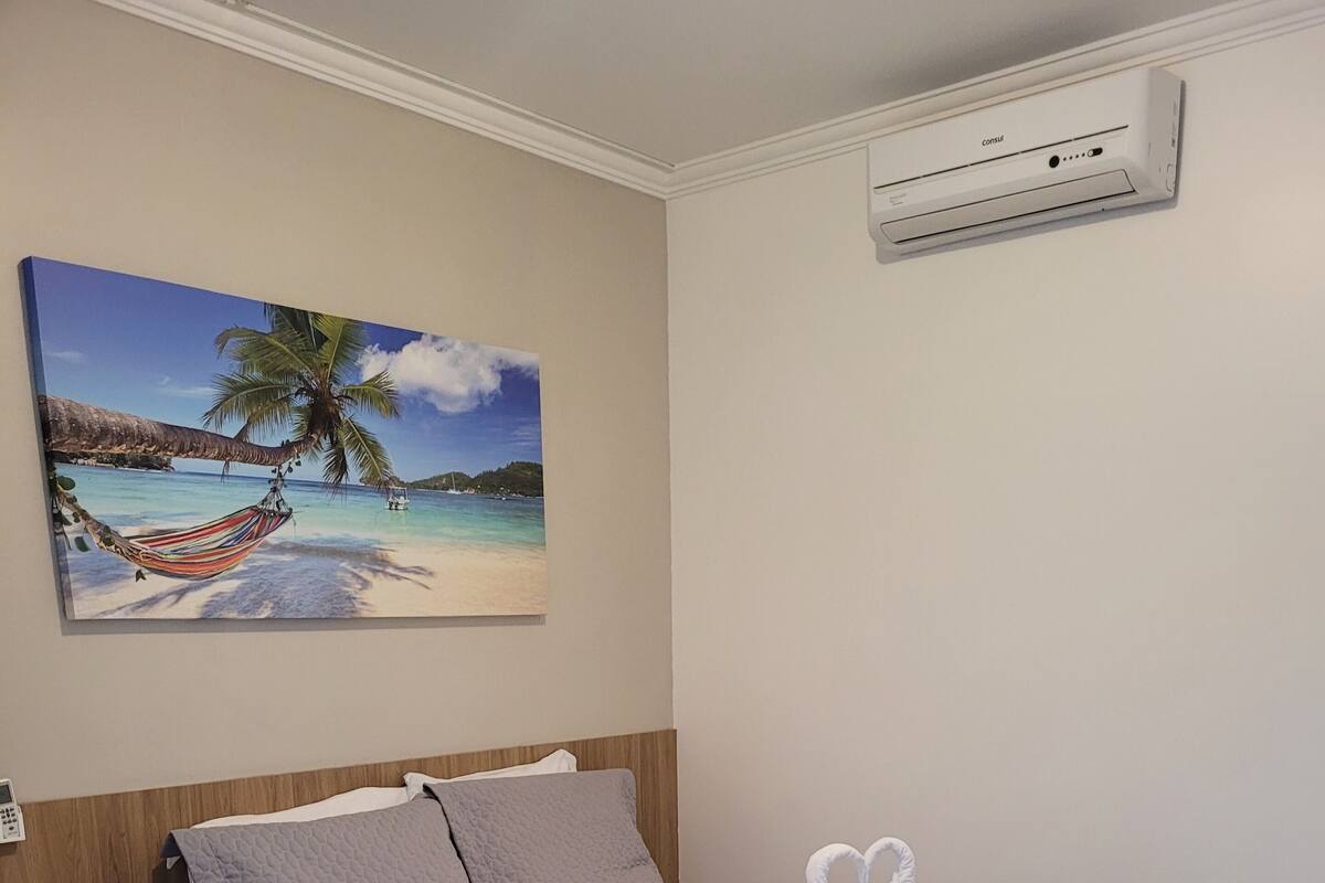 Un lit bien fait, recouvert d’une couette blanche et grise, une serviette en forme de cœur posée sur le lit et un tableau représentant une scène de plage accroché au mur.