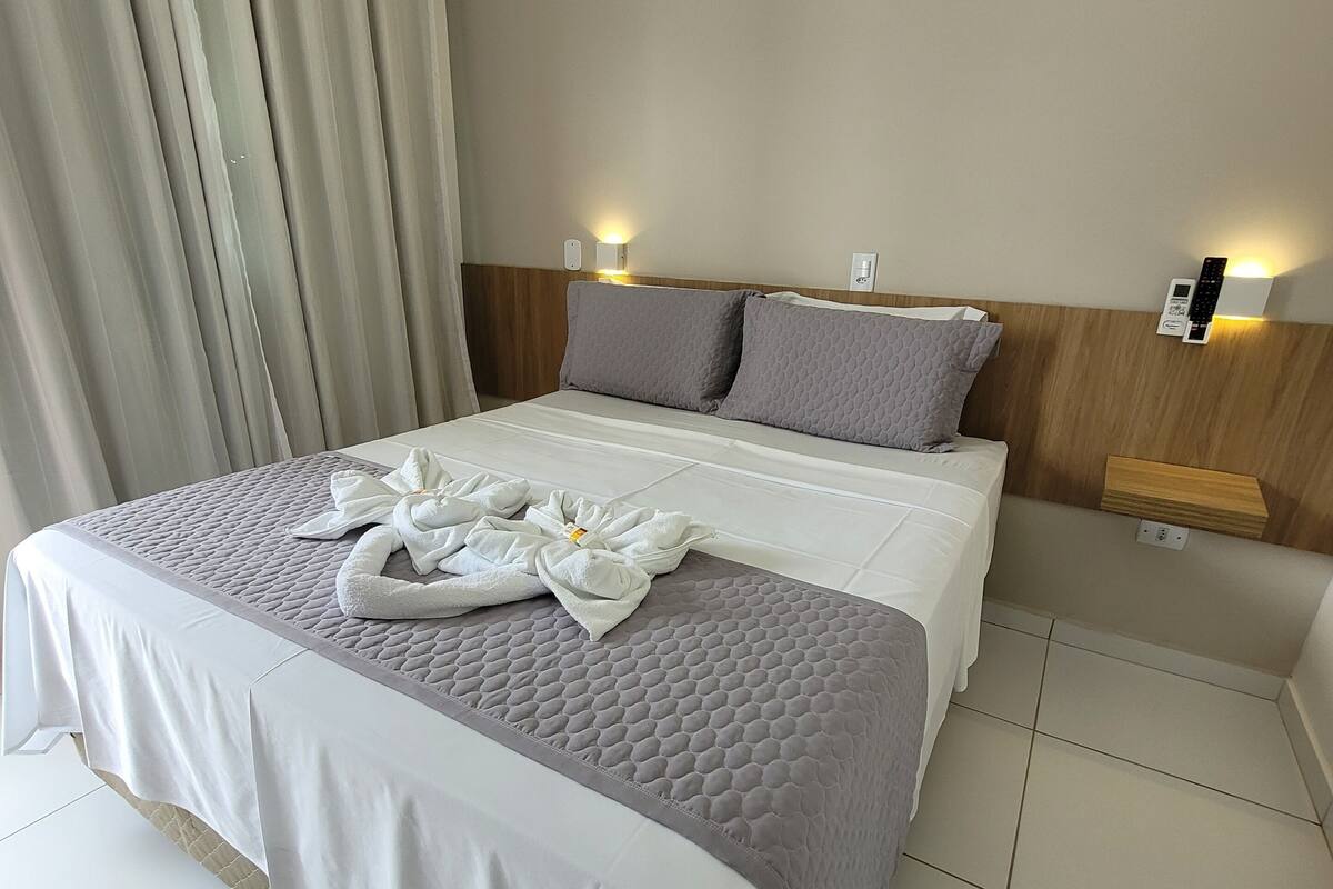 Une chambre d’hôtel avec un grand lit, deux oreillers, une table de chevet avec une lampe et un rideau.