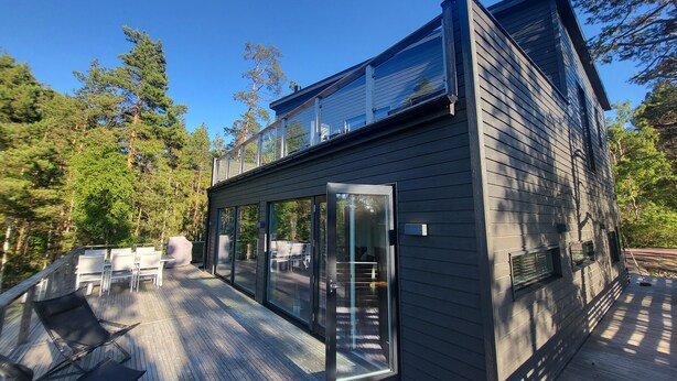 A modern two-story cabin with a deck, large glass doors, and a view of the surrounding forest. - Seaside villa in the Nordic Archipelago (Geta)