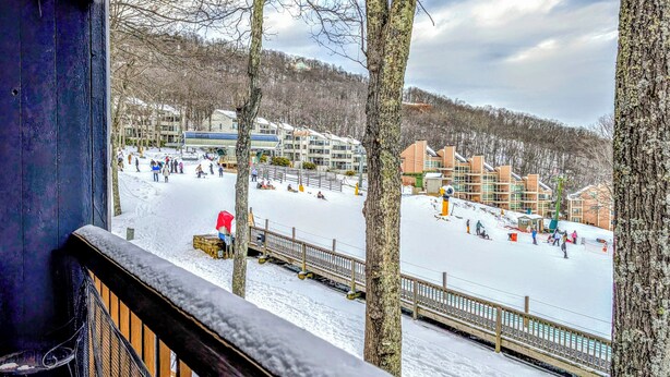 A snow-covered outdoor area with a wooden deck and railings, surrounded by buildings and trees. - Ski-In Ski-Out Wintergreen Condo w/ Balcony (Wintergreen Resort)