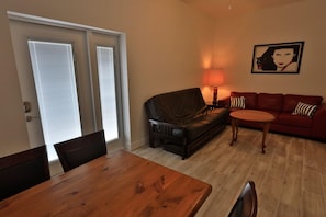 A living room with a wooden dining table, a red sofa, and a black leather couch. There is a lamp on the sofa and a framed picture on the wall.