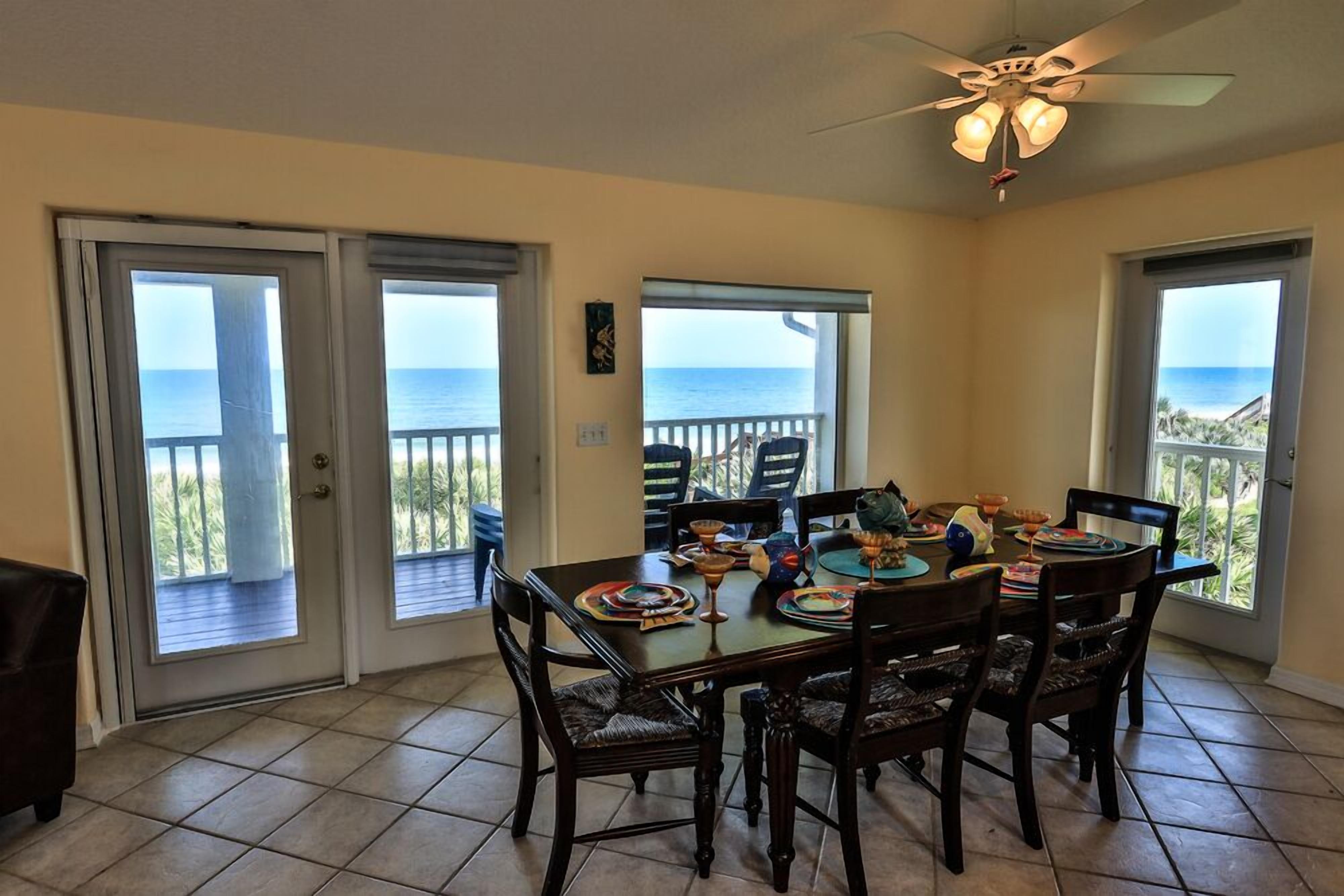 A dining area with a table set for four, surrounded by chairs, and large windows offering a view of the ocean.