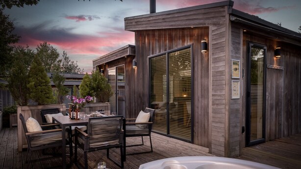 A wooden deck with a hot tub, outdoor seating, and a wooden structure with large windows. - The Retreat, Strawberryfield Park -  a lodge that sleeps 4 guests  in 2 bedrooms (Cheddar)