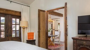Ein Schlafzimmer mit einem Bett, einer Holztür, einer Lampe, einem Fernseher und Blick in einen weiteren Raum.