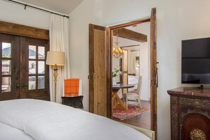 Quarto com cama, porta de madeira, luminária, televisão e vista para outro cômodo.